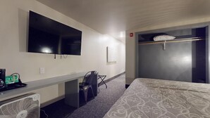 A hotel room with a bed, a desk with a chair, a television mounted on the wall, and a closet with a hanging bag.
