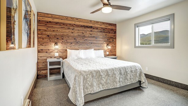 Une chambre à coucher avec une tête de lit en bois, un lit recouvert d’une couvre-lit blanche, un ventilateur de plafond, des lampes de chevet et une fenêtre donnant sur des collines.
