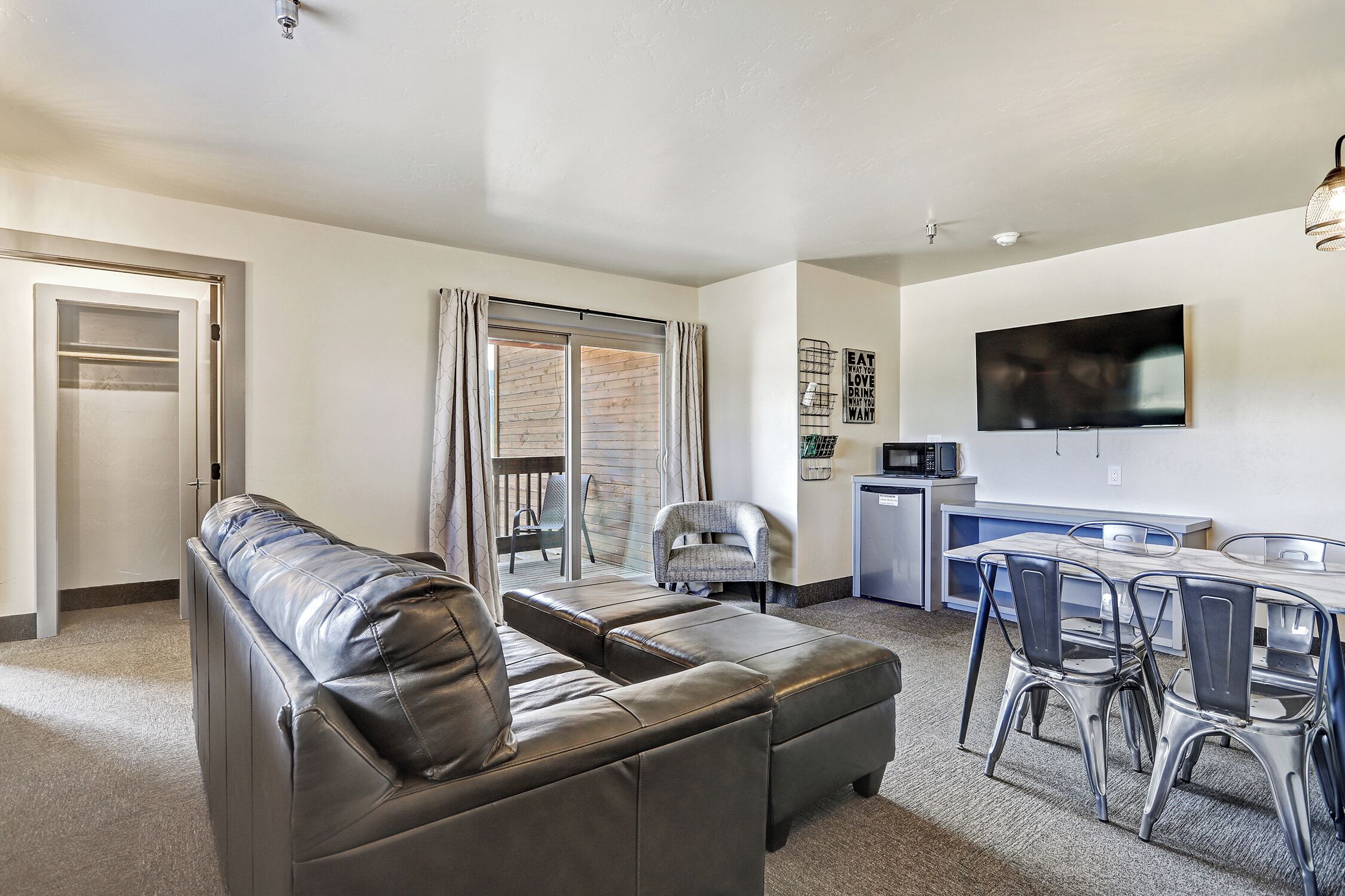 A modern living room with a leather sofa, a dining area with metal chairs, a flat-screen TV, and a kitchen area.