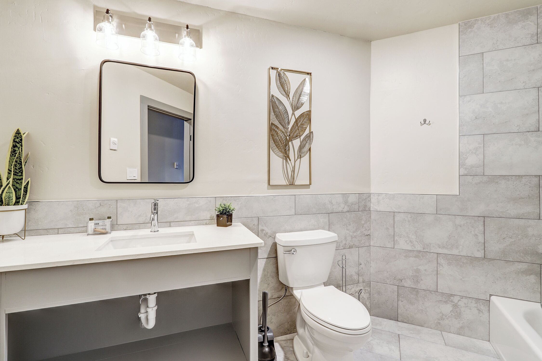 A modern bathroom with a white sink, a toilet, and a bathtub. There is a mirror above the sink and a decorative leaf wall art piece.