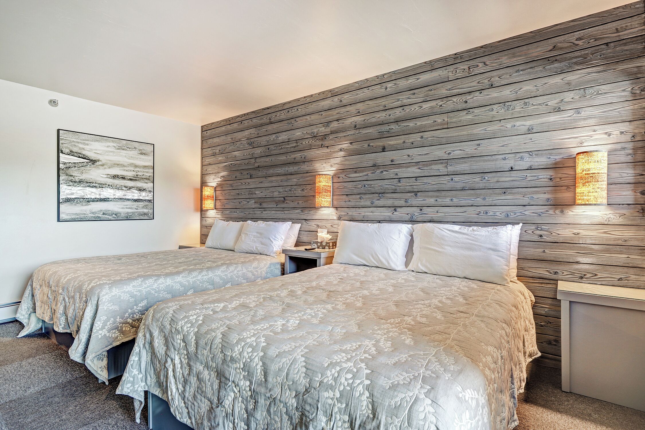 A hotel room with two beds, a wooden headboard, wall-mounted lights, and a framed picture.