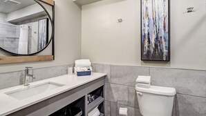 Ein modernes Badezimmer mit einem weißen Waschbecken, einem Spiegel mit Holzrahmen, einer Toilette und einem großen, abstrakten Gemälde an der Wand.