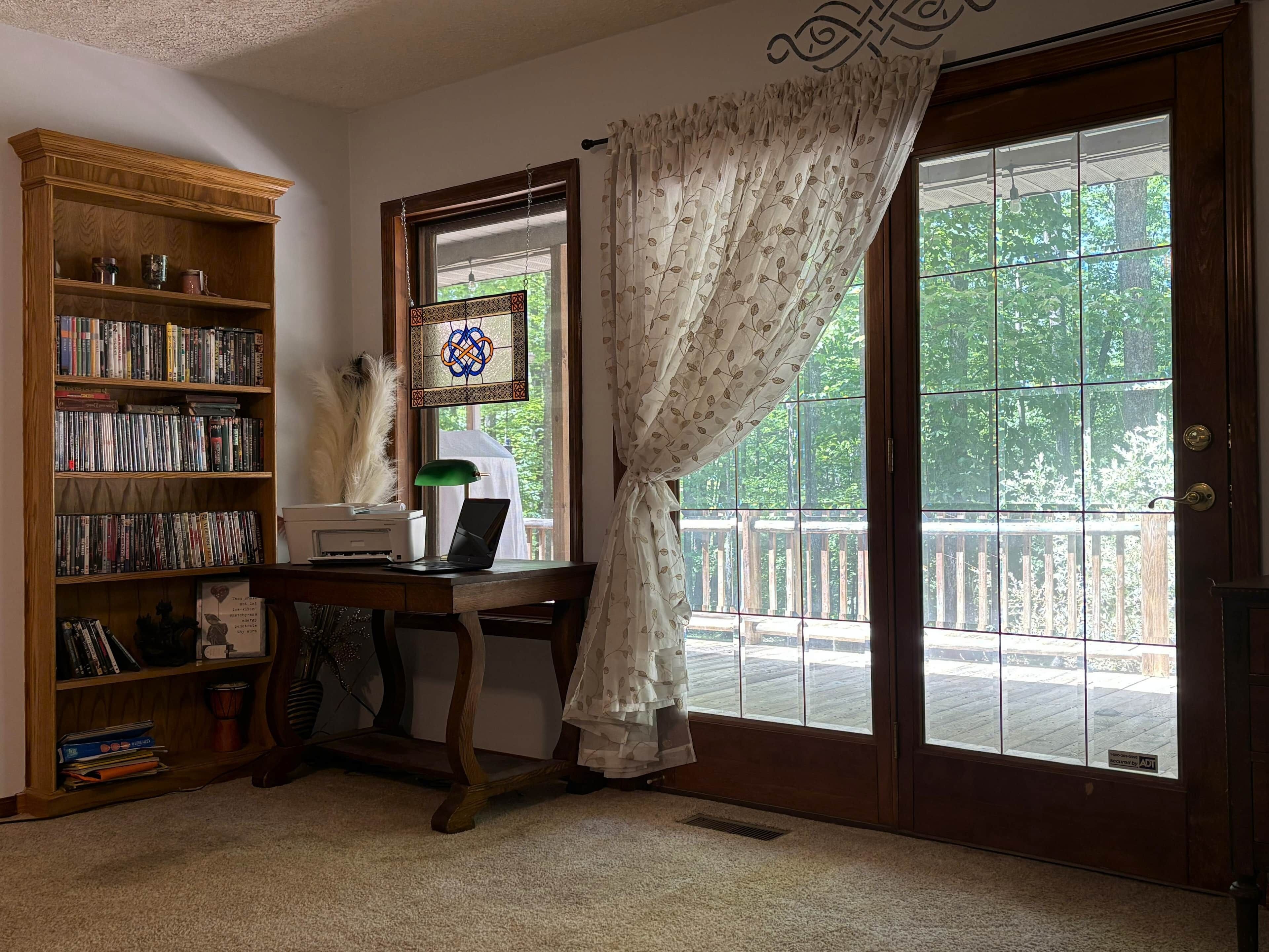 Una habitación con una estantería de madera, un escritorio con un ordenador portátil, un ventanal decorativo y vistas a un balcón.