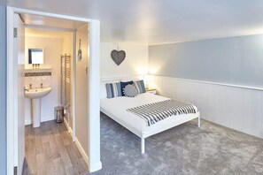 A bedroom with a white bed, a bedside table, a wall-mounted heart decoration, and a bathroom visible through an open door.