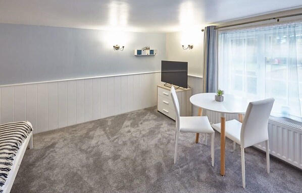 A room with a bed, a desk with a computer, a chair, a small table with a plant, and a window with curtains.