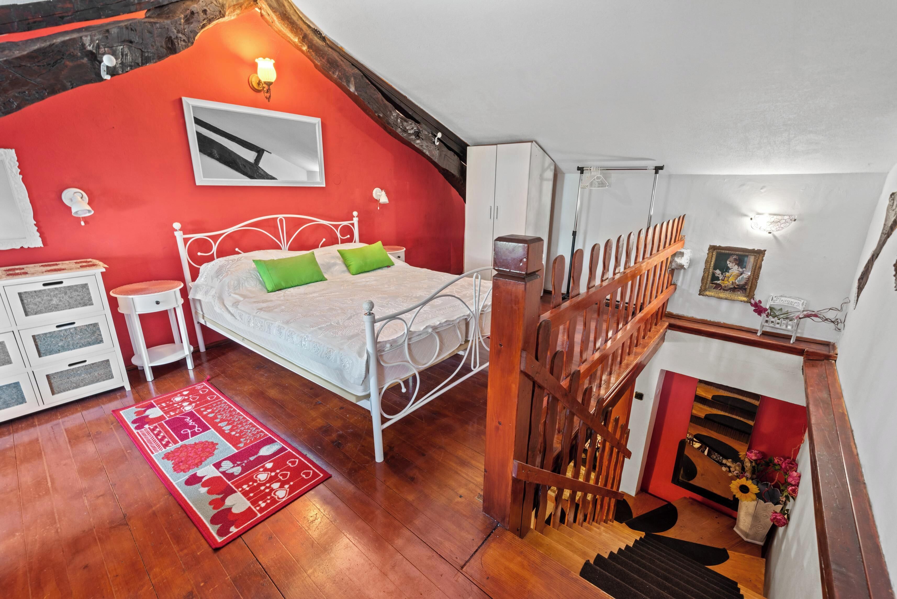 A bedroom with a bed, a nightstand, a dresser, a red wall, a wooden staircase, and a small altar with flowers.