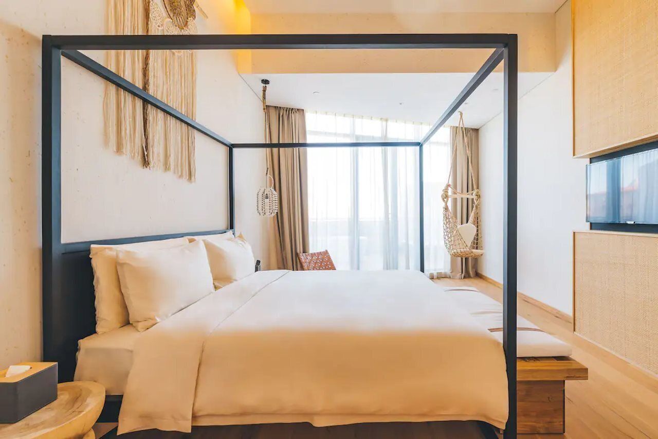 Una habitación de hotel moderna con una cama con dosel, una mesita de noche de madera, una mesita auxiliar y un gran ventanal con cortinas.
