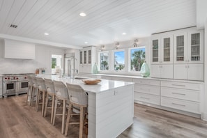 A modern kitchen with white cabinetry, a central island with bar stools, a built-in oven, and a view of the outside through large windows.