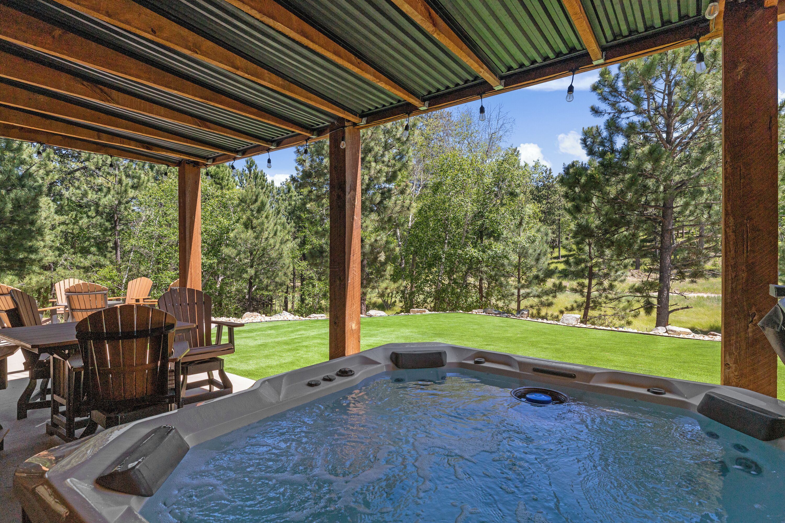 A hot tub with a wooden deck and chairs, surrounded by a grassy yard and trees.