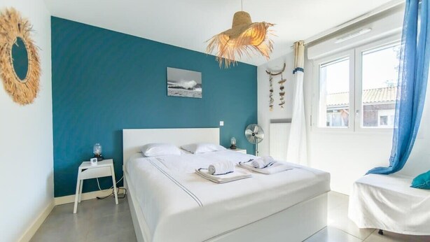 A bedroom with a bed, a bedside table, a wall-mounted fan, and a window with curtains. - Appartment Christine in Lège city for 5 persons (Lège-Cap-Ferret)