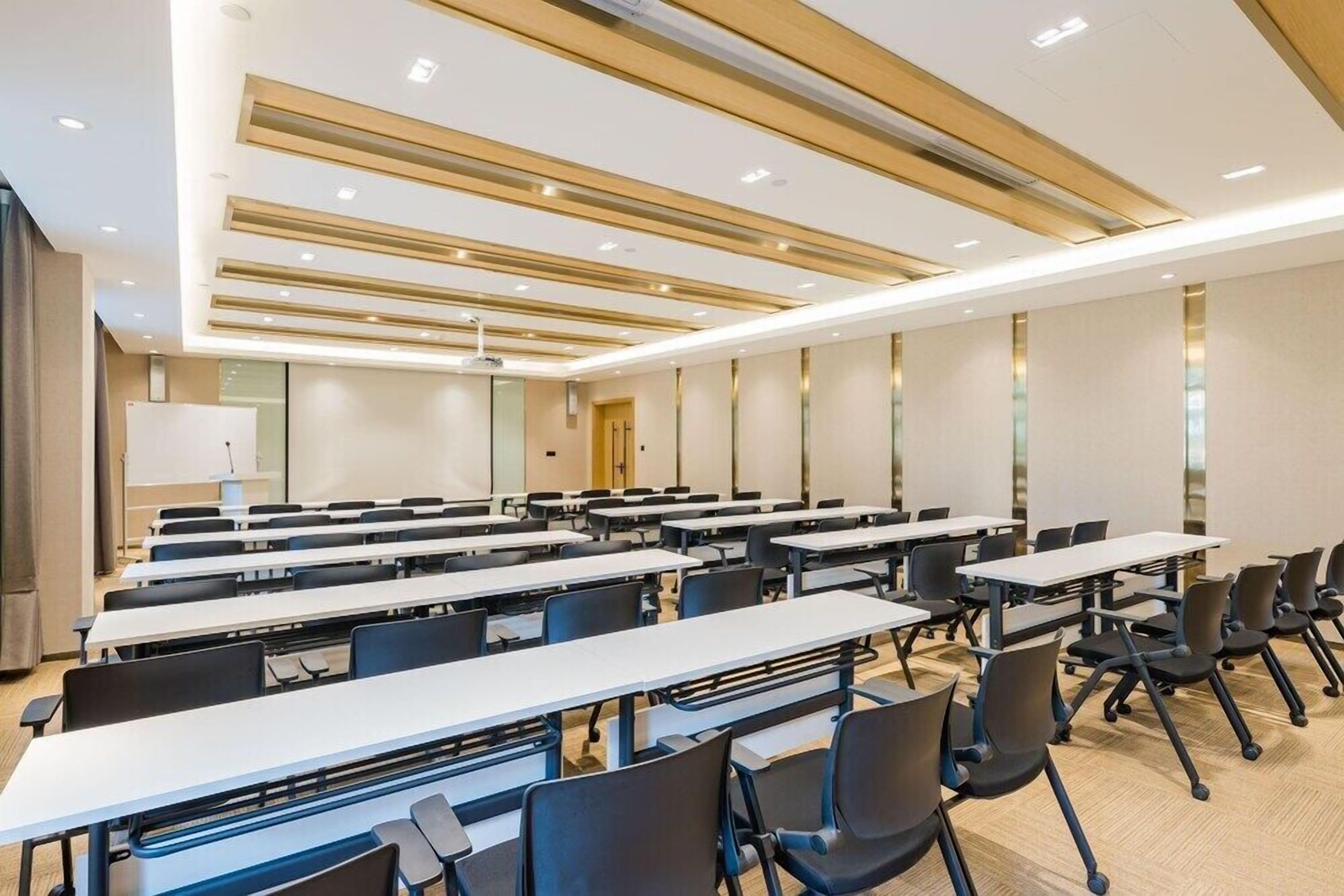 A conference room with rows of tables and chairs, a modern ceiling design, and a neutral color scheme.