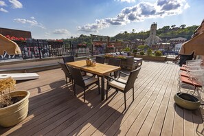 Eine Holzterrasse mit Tisch und Stühlen, Blumentöpfen und Blick auf eine Kirche sowie Gebäude im Hintergrund.