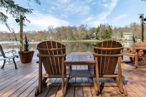 Eine Holzterrasse mit zwei Adirondack-Stühlen, die auf einen See ausgerichtet sind, einem kleinen Tisch und einem Blumentopf.