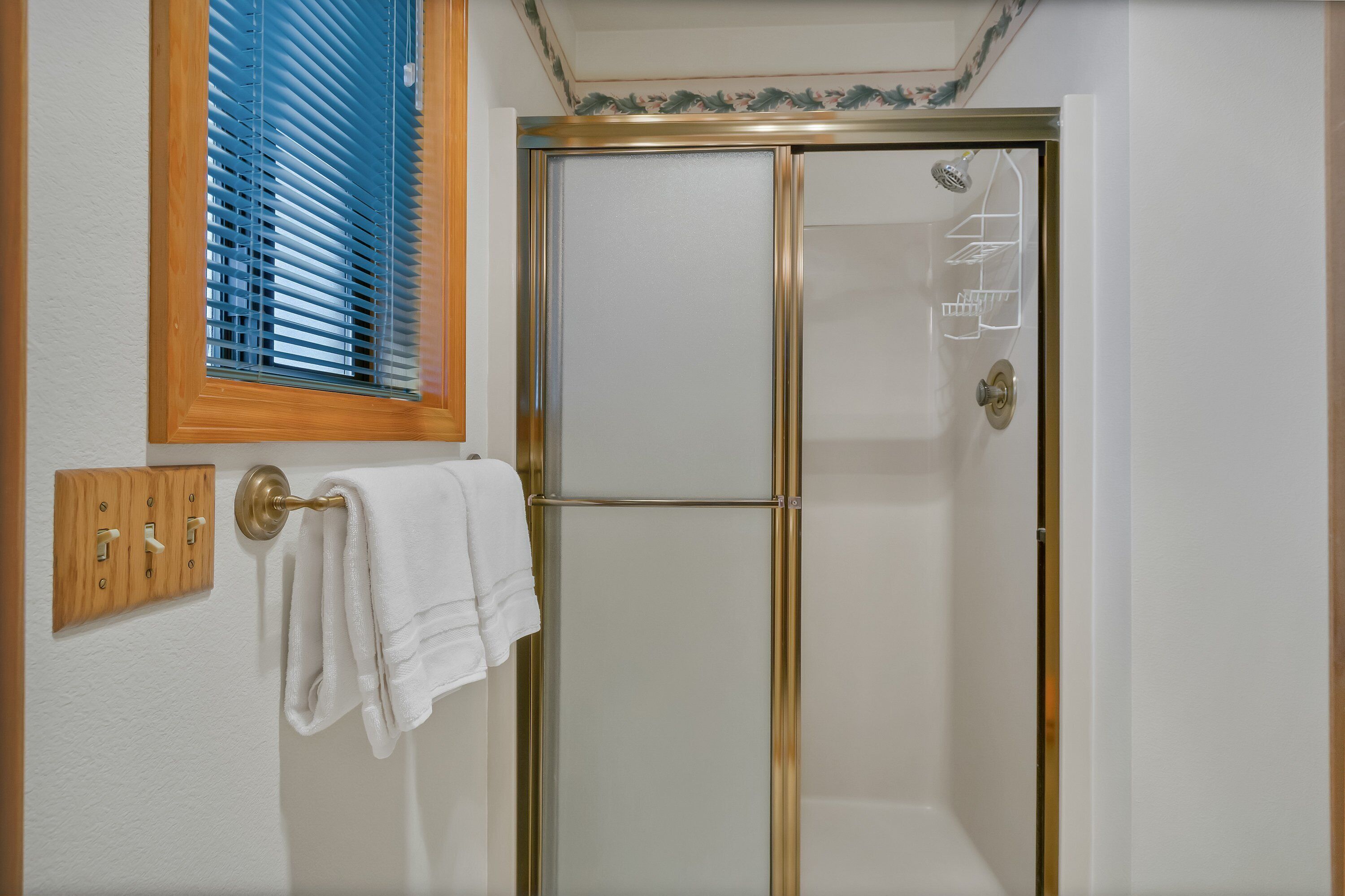 A bathroom with a glass shower door, a towel rack with white towels, and a window with blinds.