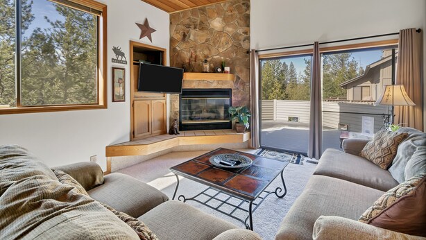 A cozy living room with a stone fireplace, a flat-screen TV, and a large window offering a view of the outdoors.