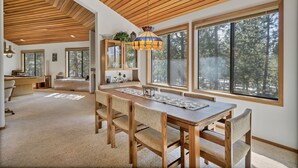 A spacious dining area with a wooden table and chairs, large windows with a forest view, and a ceiling with wooden slats.