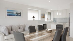 Eine moderne Küche mit weißen Schränken, ein Essbereich mit Holztisch und -stühlen sowie ein Wohnbereich mit einer Couch und Kissen.