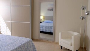 Um quarto com uma cama, uma poltrona branca, um espelho grande e um candeeiro fixado na parede.