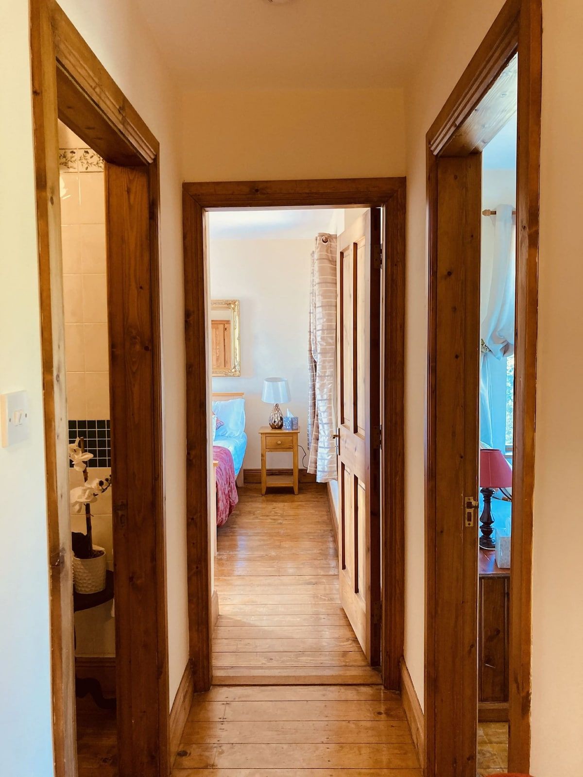 A narrow wooden hallway leading to a bedroom with a bed, a nightstand, and a lamp.