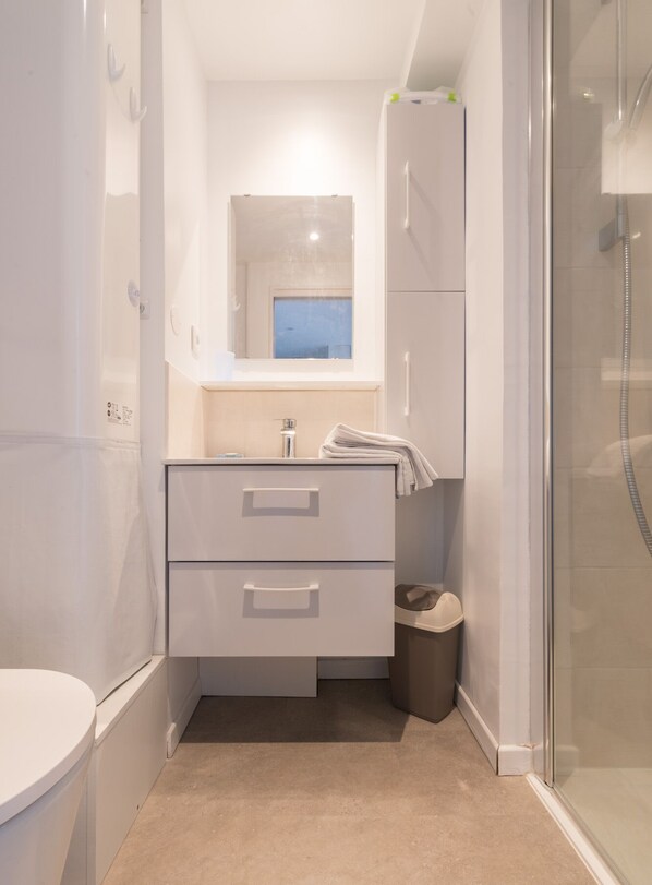 Ein modernes Badezimmer mit Toilette, einem Waschbecken mit Spiegel darüber, einer Dusche und einem Mülleimer.