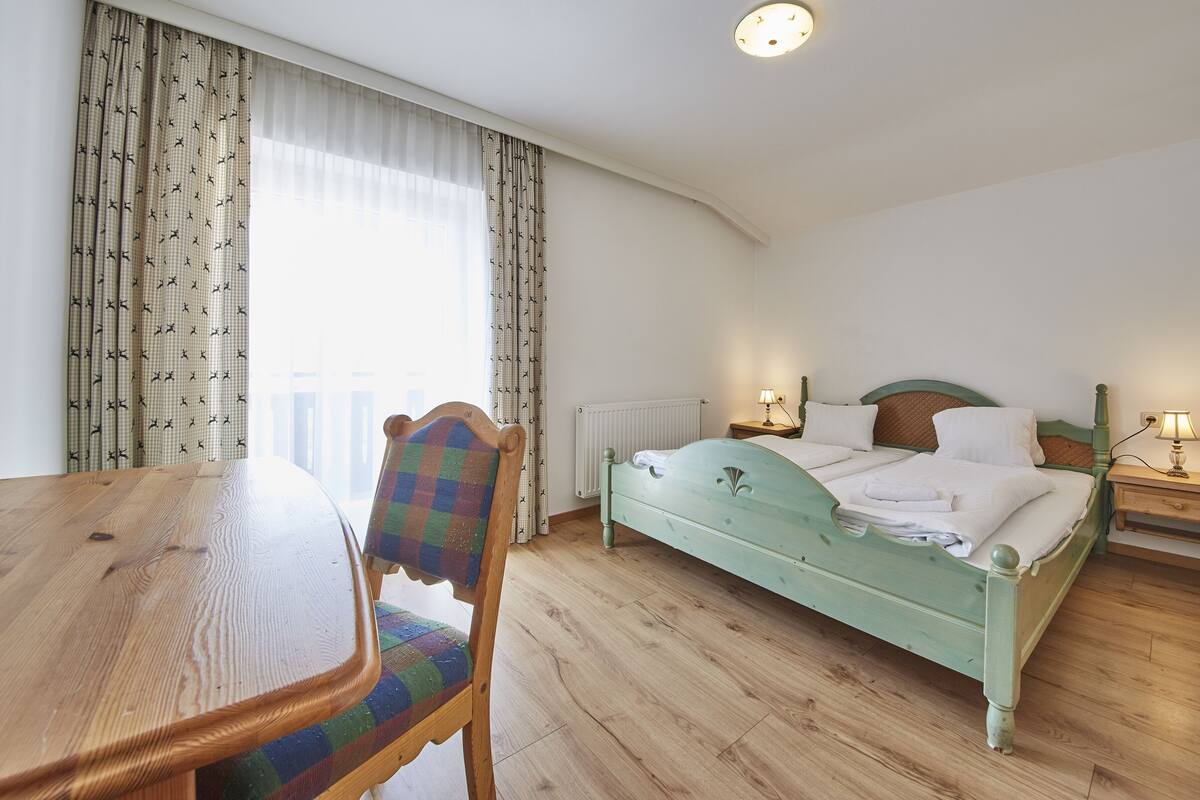 Ein Schlafzimmer mit einem hölzernen Bett, einem Stuhl, einem Tisch und einem Fenster mit Vorhängen.
