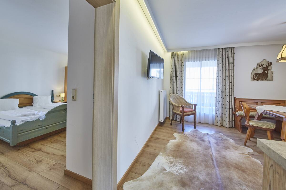 Ein Hotelzimmer mit Bett, Sessel, Esstisch, Fernseher und einem großen Fenster mit Vorhängen.