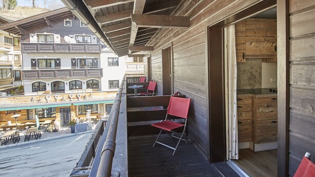 A wooden balcony with red chairs, a view of a street, and a building with multiple windows in the background. - TownHouse Appartements (Saalbach-Hinterglemm)
