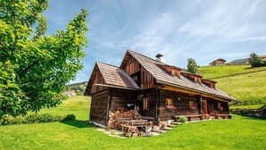 Ein rustikales Holzhaus mit Satteldach, einer Veranda und einem kleinen Garten mit Schubkarre und Holzscheiten.