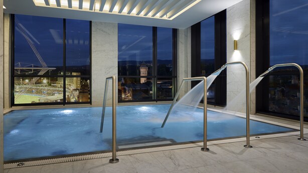 A modern indoor swimming pool with clear glass railings, large windows offering city views, and a contemporary ceiling design.