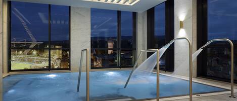 A modern indoor swimming pool with clear glass railings, large windows offering city views, and a contemporary ceiling design.