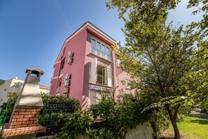 Un bâtiment rose doté d’un balcon, une structure en pierre et un arbre au premier plan.