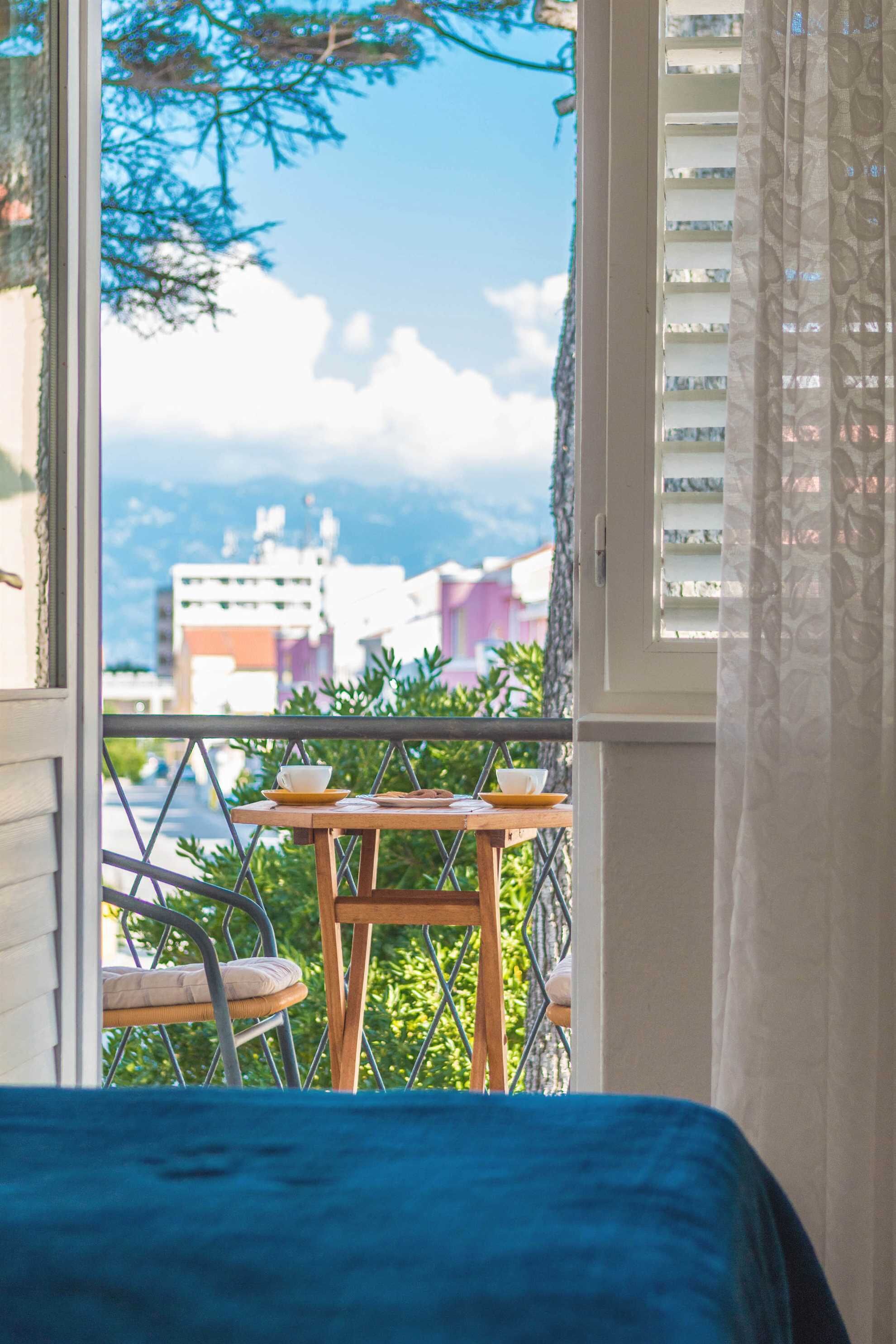 A balcony with a table and chairs, a view of buildings and mountains, and a window with blinds.