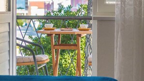 Un balcón con mesa y sillas, vista a edificios y montañas, y una ventana con persianas.