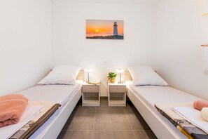 Ein Doppelzimmer mit zwei Betten, einem Nachttisch mit Lampen, einem kleinen Tisch mit einer Pflanze und einem Gemälde eines Leuchtturms an der Wand.
