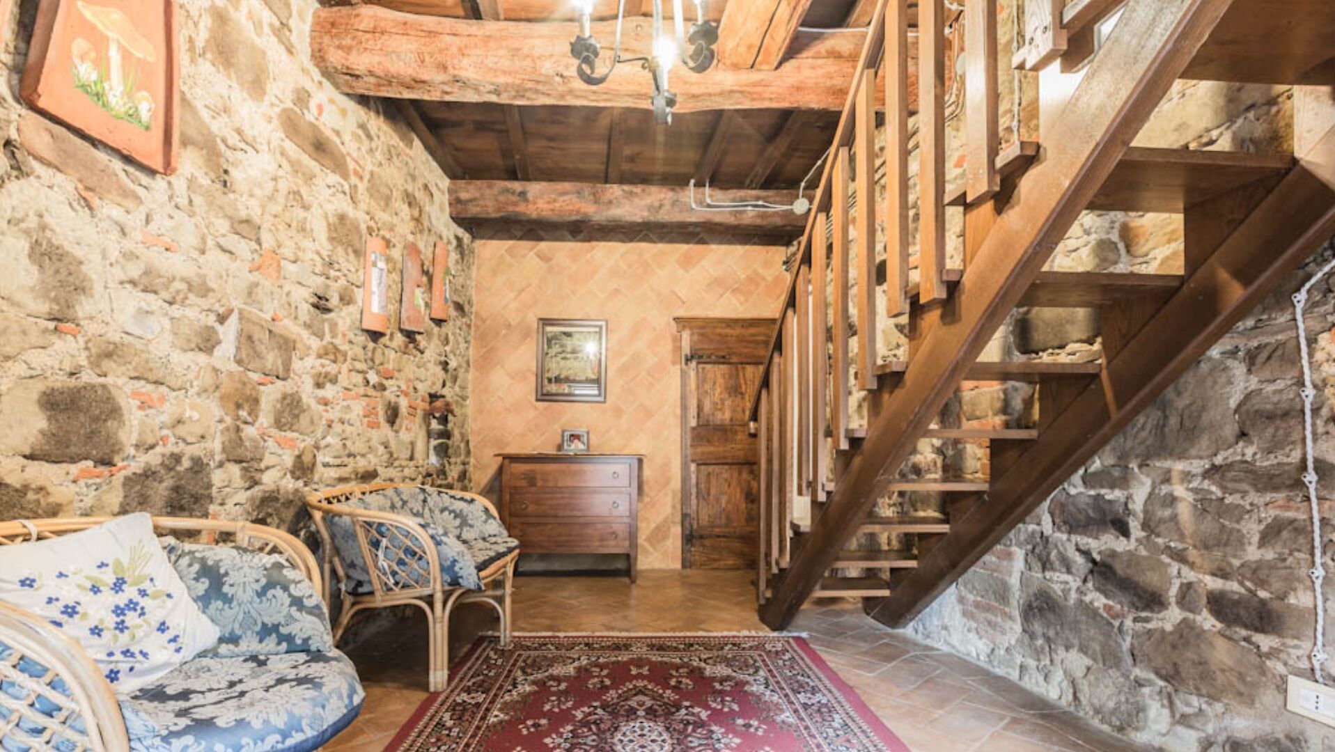 A rustic room with stone walls, a wooden staircase, a wooden chest of drawers, and a patterned rug.