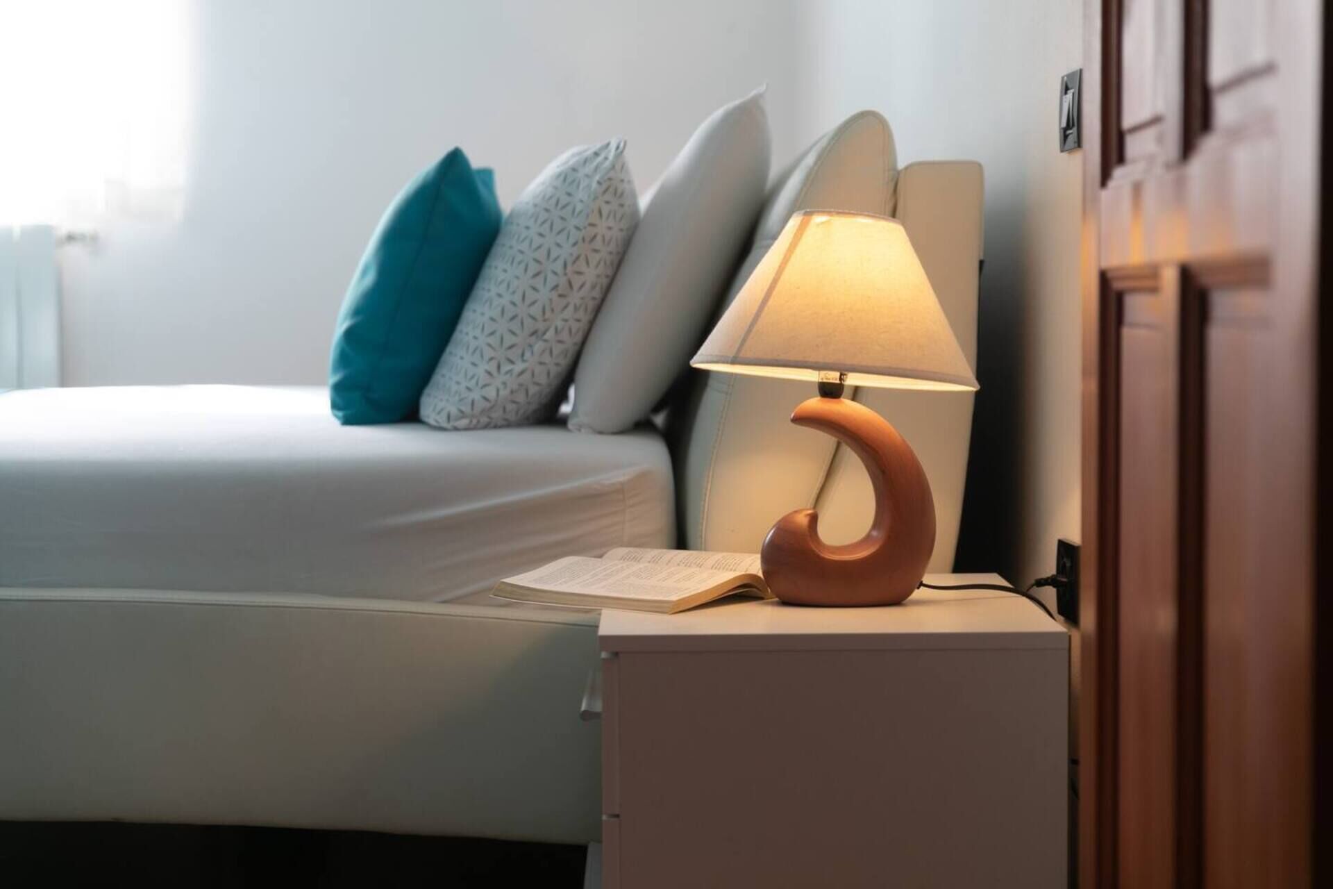 A neatly made bed with a white comforter and a teal pillow. A wooden nightstand with a lamp is beside the bed.