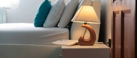 A neatly made bed with a white comforter and a teal pillow. A wooden nightstand with a lamp is beside the bed.