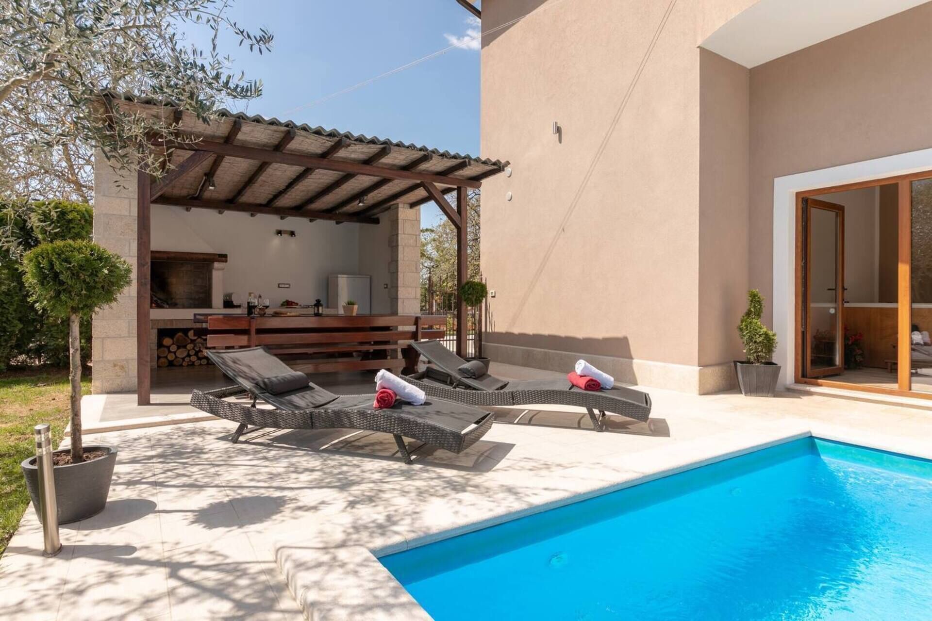 A poolside area with two lounge chairs, a pergola, and a barbecue setup.