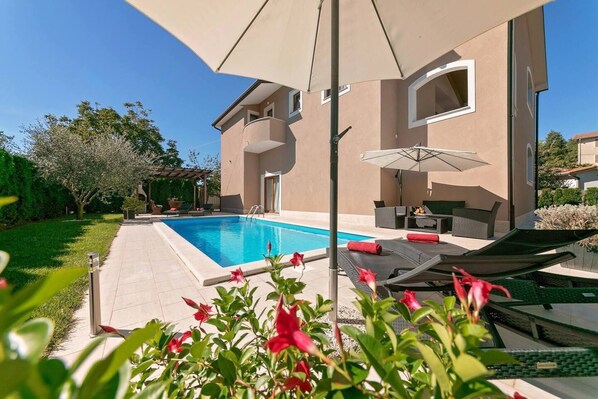A swimming pool with a sun umbrella, outdoor seating, and a garden.