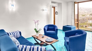 Una habitación con un sofá azul, un sillón azul, una mesa de madera con un plato azul y blanco, una planta en maceta y vistas a un balcón con barandilla.