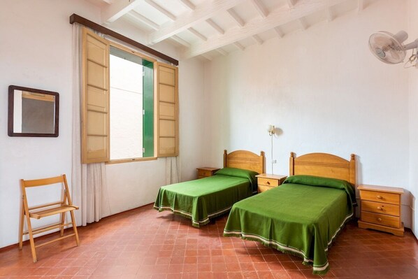 A room with two single beds, a wooden chair, a small table, and a window with open shutters.