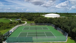 A tennis court with a net, surrounded by a fence, and a large white building in the background.