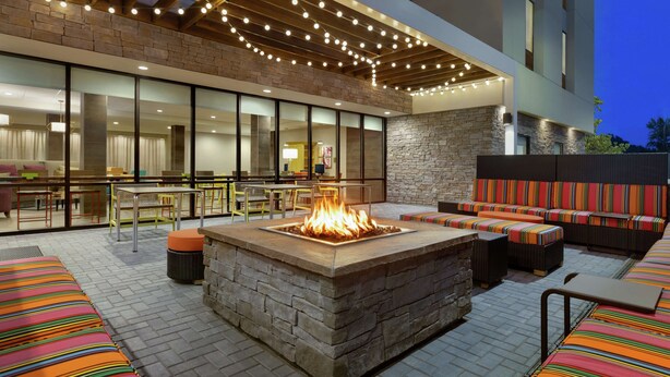 An outdoor patio area with a stone fire pit, seating with colorful cushions, and a dining area with tables and chairs. - Home2 Suites by Hilton New Brunswick, NJ (New Brunswick)