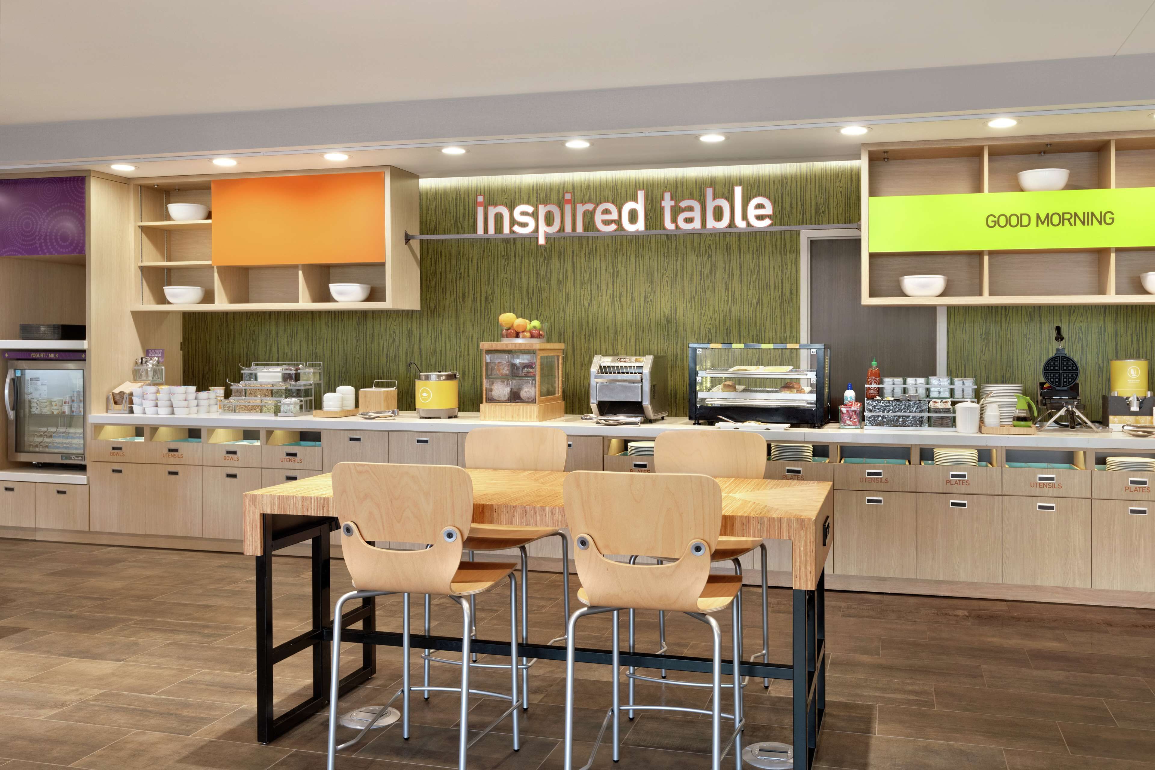 A modern cafe with a wooden table and chairs, a counter with various items, and a sign that reads "inspired table".
