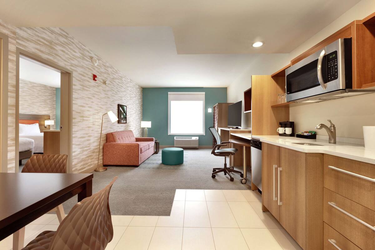 A modern hotel room with a kitchenette, a dining area, a living area with a sofa and a lamp, and a large window providing natural light.