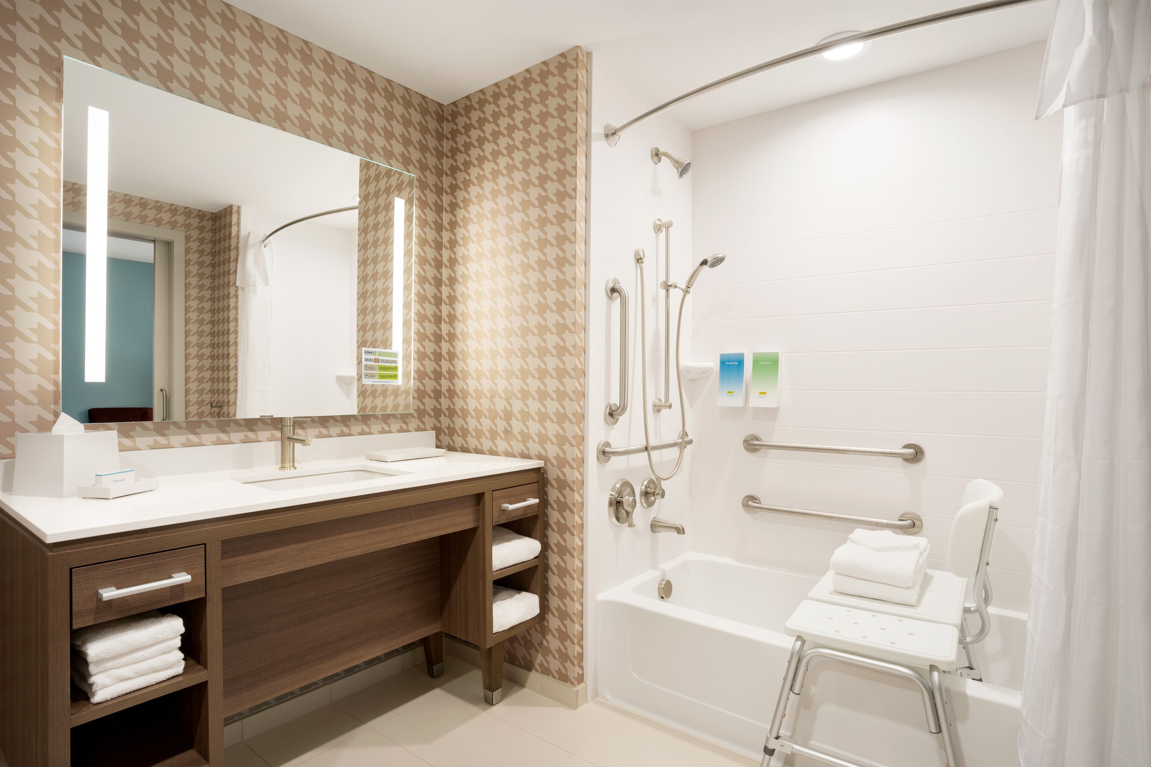 Un bagno moderno con un mobile bagno in legno, un ampio specchio, una vasca da bagno con una sedia a doccia e una barra di sostegno.