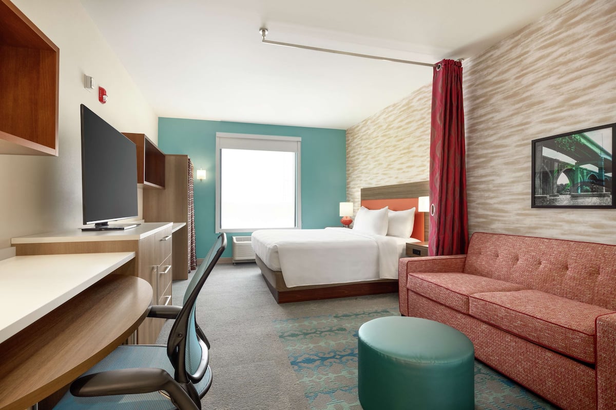 A hotel room with a bed, a desk with a computer, a TV, a sofa, and a window with curtains.