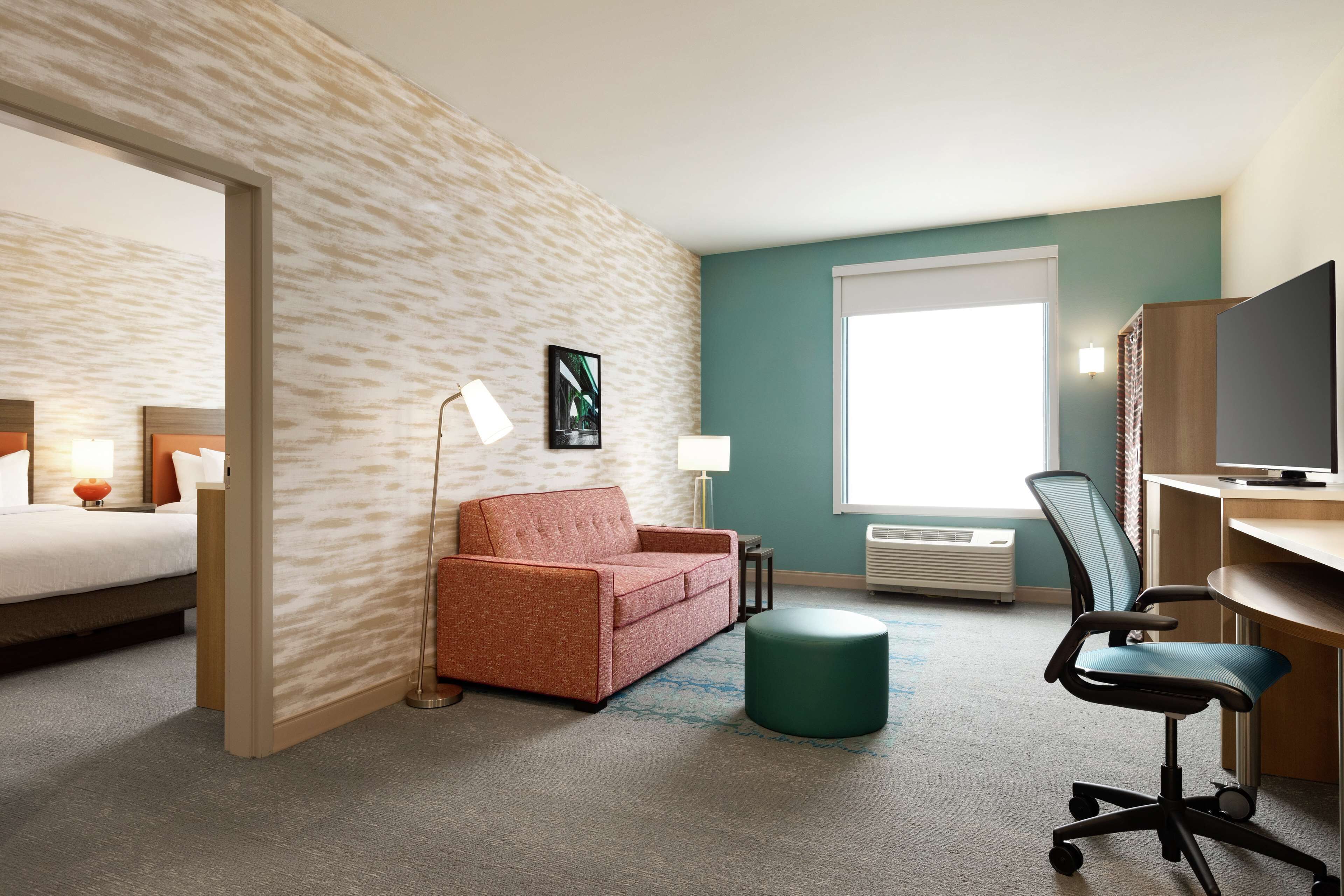 A hotel room with a bed, a red sofa, a green ottoman, a desk with a chair, and a television.