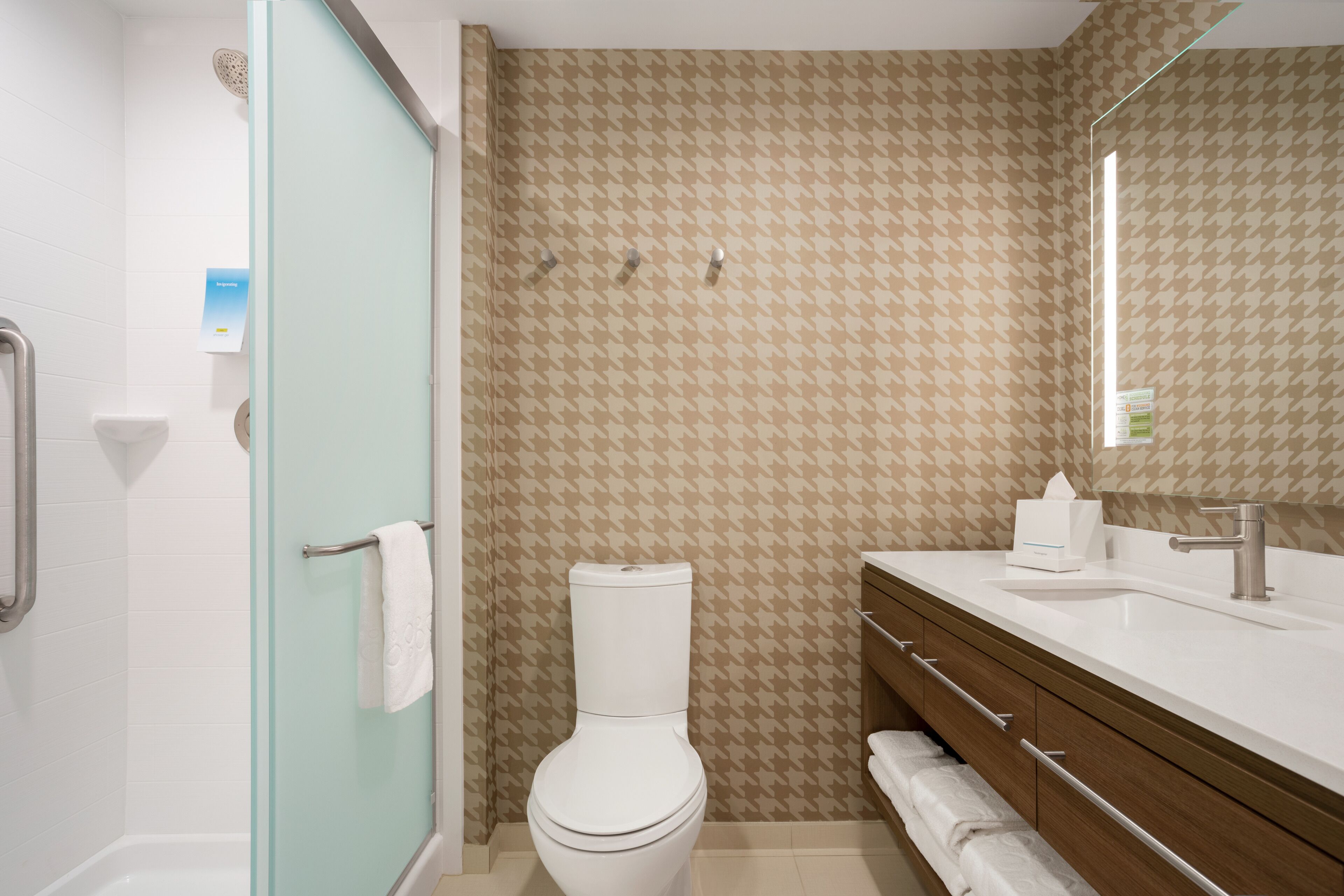 A bathroom with a white toilet, a glass shower enclosure, a wooden vanity with a mirror, and a patterned wall.
