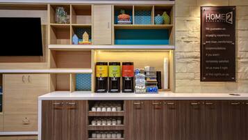 A hotel reception area with a coffee station, a TV, and a sign stating 'HOME 2 GUARANTEE'.